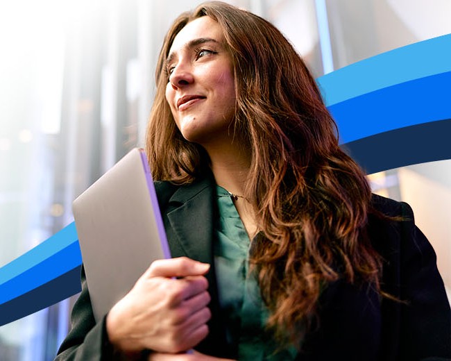 A smiling woman with long brown hair, dressed professionally, holds a laptop and looks confidently into the distance.